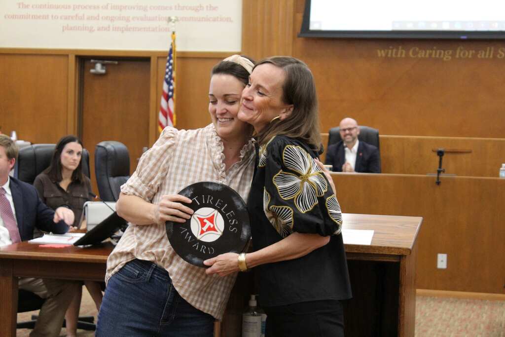 Two women hugging while holding a plaque with a red logo. A flag in the background.