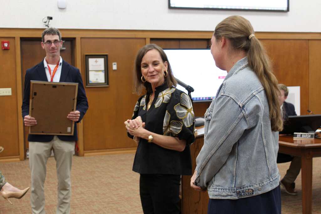 Three people, one with a clipboard, talk near a podium with a monitor and a desk.
