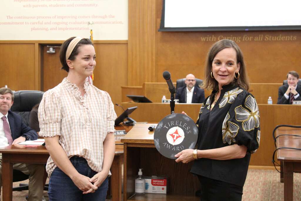 Two women holding an award plaque, standing in front of a desk with a microphone in a conference room.