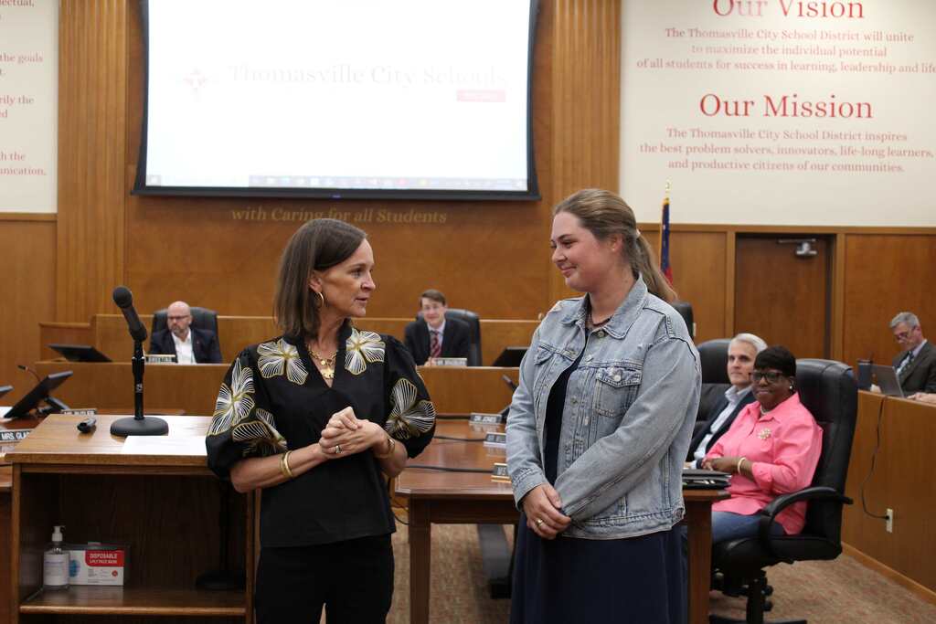 Two women stand and converse in a room with a table and chairs, a podium, and a screen.