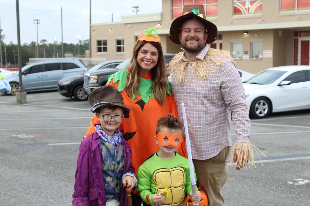 Two adults and three children in costumes stand in a parking lot with vehicles and a building in the background.