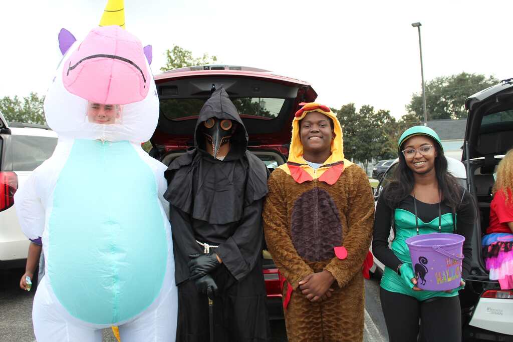 Five individuals dressed in costumes standing in a parking lot, one in an inflatable unicorn costume, one in a dark robe, one in a lion costume, and two in colorful costumes.