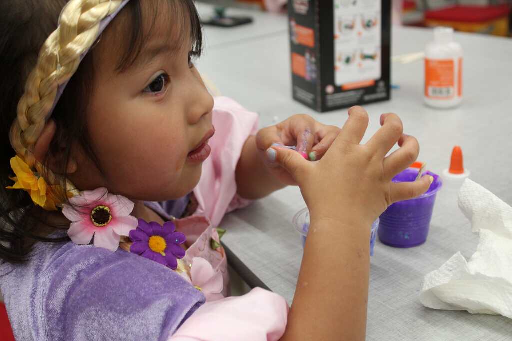 Child in purple dress and headband sitting at table with hands on paper cup and tissue.