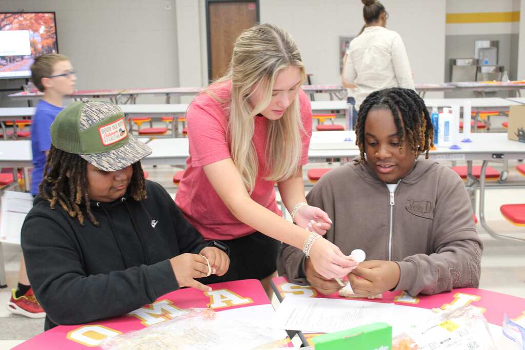 Three students collaborate at a table with food preparation items, guided by an adult wearing a pink shirt.