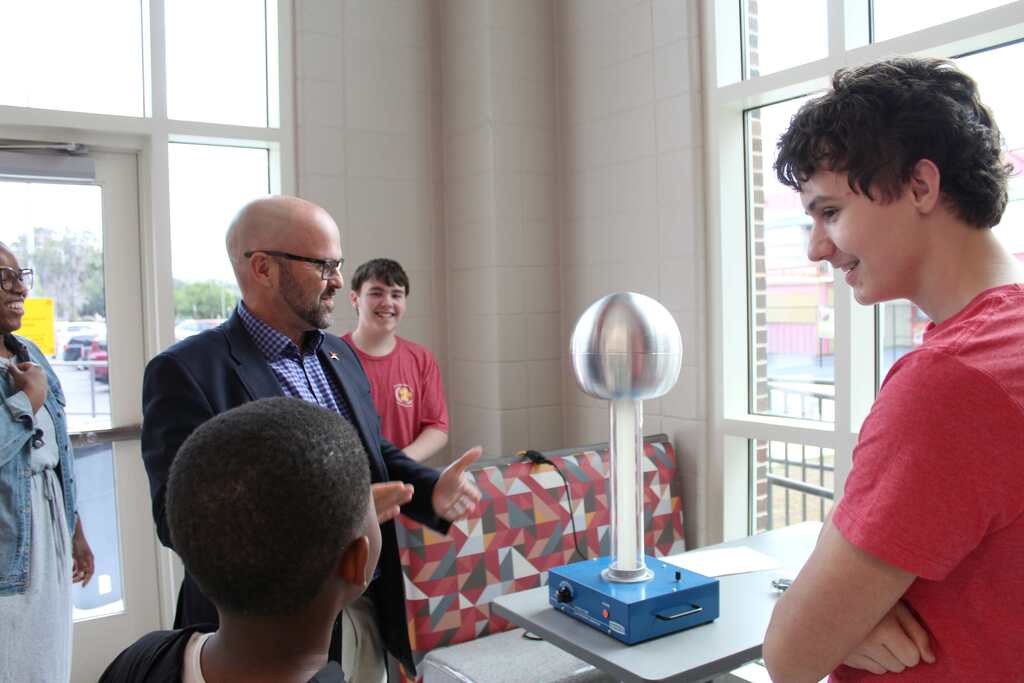 A man speaks to two students in front of a metal sphere. A boy with glasses stands in the background.
