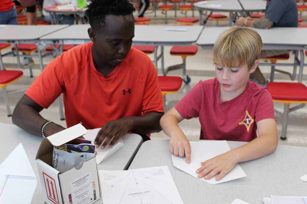 Two students working at desks; one with a box of items, the other with papers.