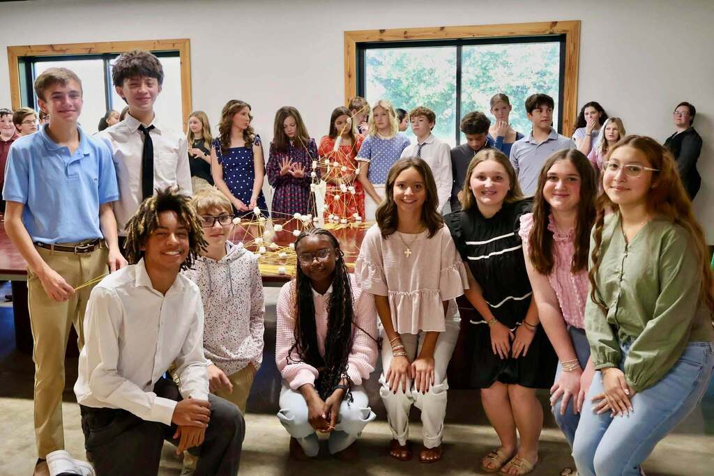 Students pose for a group photo in front of a conference table filled with spaghetti and marshmallow towers, showcasing their teamwork and creativity during the Emerging Young Leaders kickoff activity.