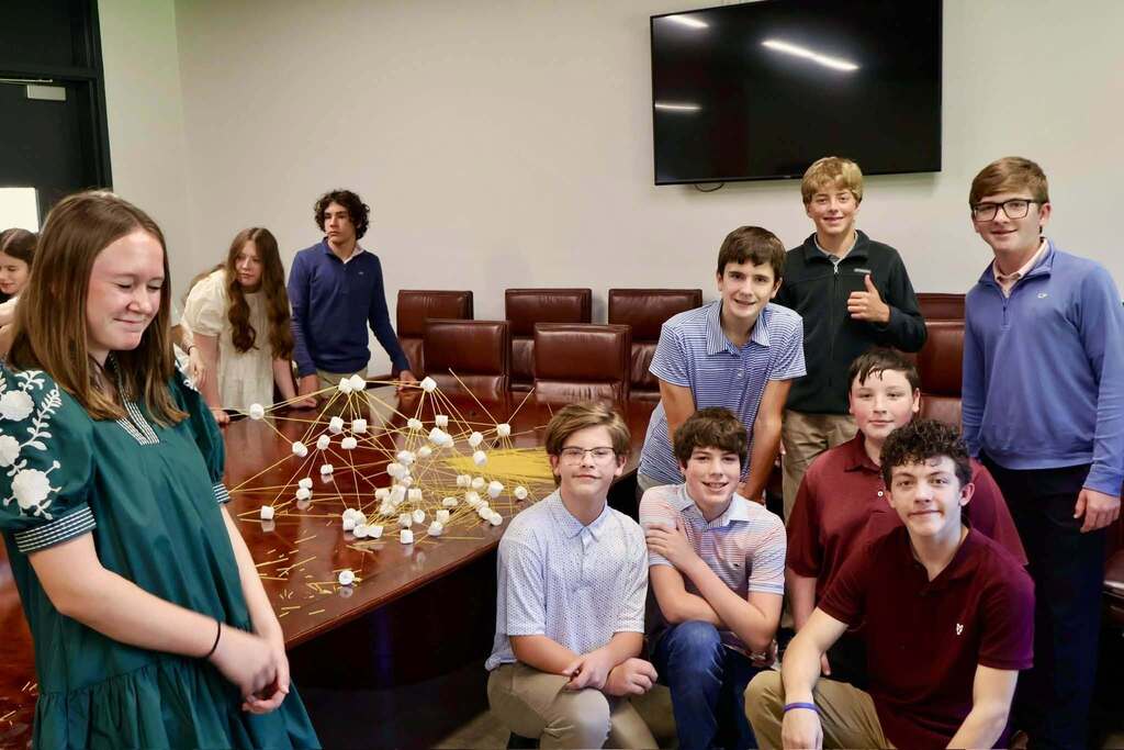 Students participating in the 2026–2027 Emerging Young Leaders Program pose around a conference table displaying a large tower made from spaghetti and marshmallows. Several students stand behind the table while others kneel or sit in front, showcasing their completed teamwork challenge during the kickoff event hosted by the Tifton-Tift County Chamber of Commerce.