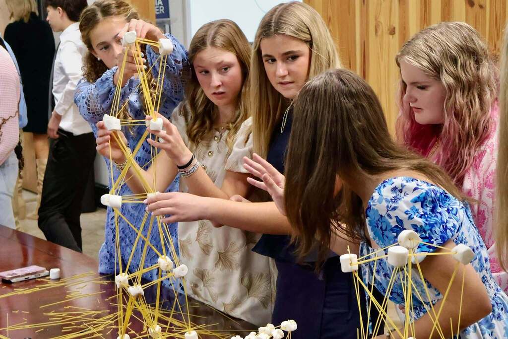 Five students work together to stabilize a tall spaghetti and marshmallow tower, carefully holding and adjusting its structure during the Emerging Young Leaders kickoff event.