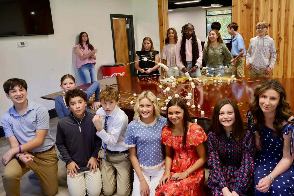 Students gather around a conference table covered with spaghetti and marshmallow structures during the Emerging Young Leaders kickoff event. Several students pose in the foreground while others stand behind their creations.