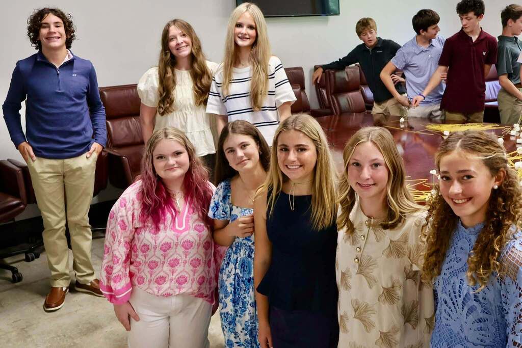 A group of students smiles for a photo inside the Chamber meeting room while other participants continue working on spaghetti and marshmallow towers in the background.