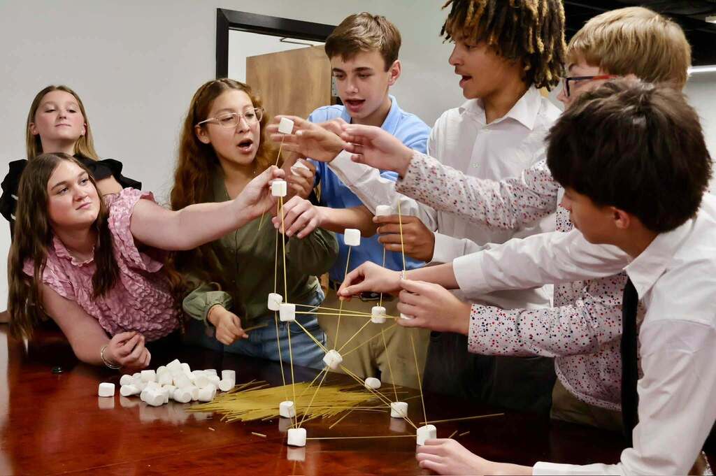 A group of students collaborates around a conference table, carefully balancing spaghetti strands and marshmallows to build a tower during the Emerging Young Leaders teamwork challenge.