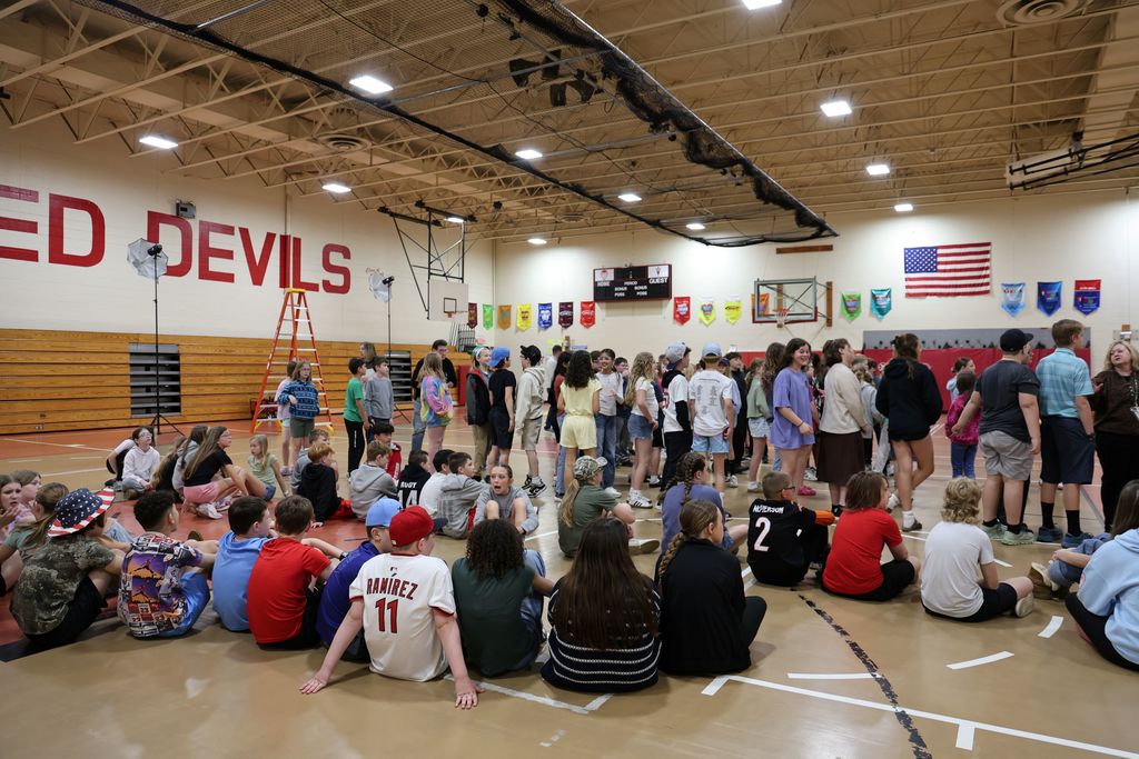 Fifth graders on the floor in the gym and some standing as they listen to the directions.