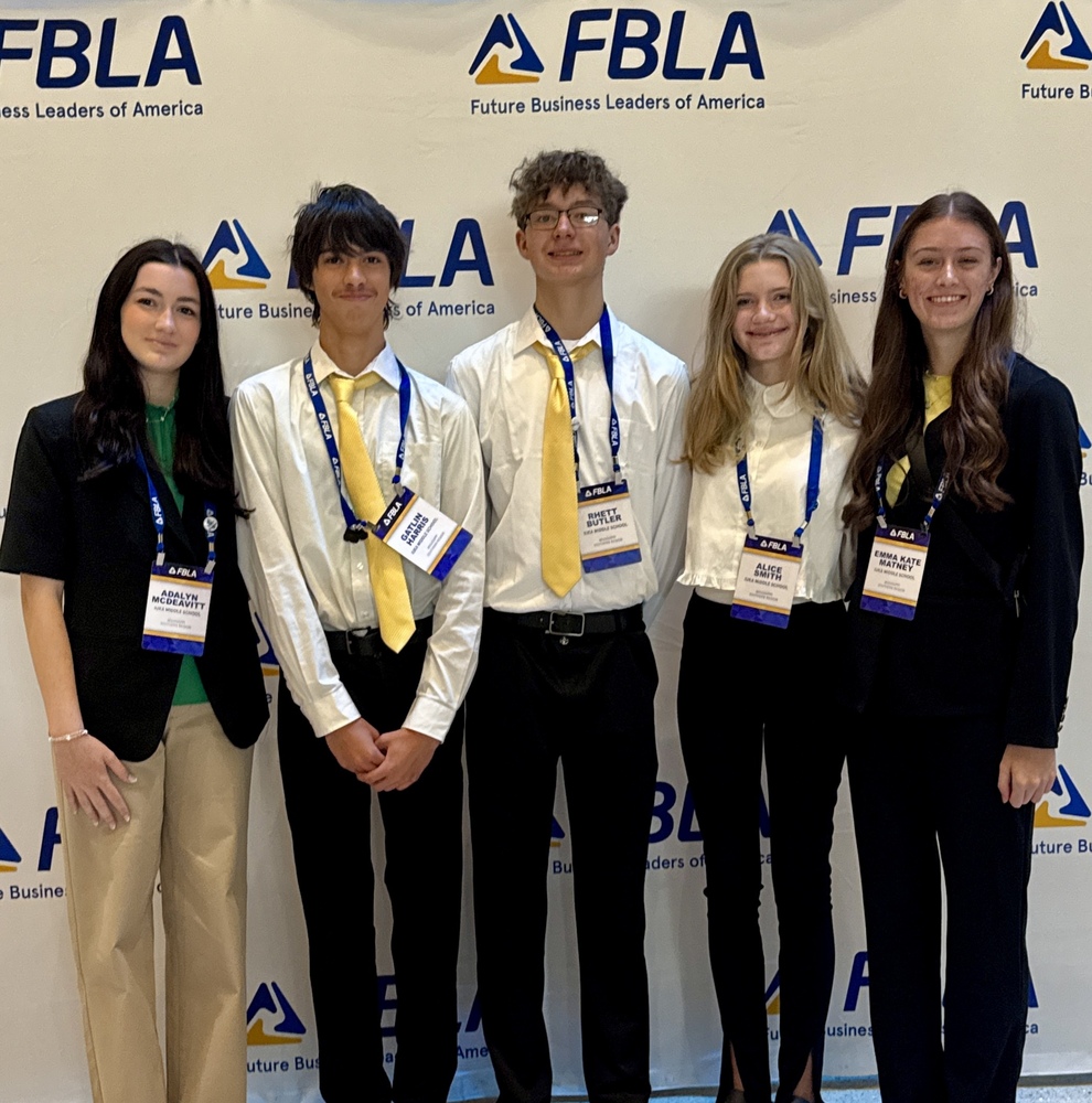 Five middle school students from the Iuka Middle School FBLA chapter stand in a line posing together at the FBLA National Leadership Conference. They are positioned in front of a white backdrop repeatedly printed with the blue and yellow Future Business Leaders of America (FBLA) logo. From left to right: a girl wearing a black blazer, green shirt, and tan pants; a boy in a white dress shirt, yellow tie, and black slacks; a taller boy wearing a white dress shirt, yellow tie, glasses, and black slacks; a girl wearing a white ruffled blouse and black slacks; and a girl wearing a black pantsuit with a yellow top underneath. Each student wears a blue FBLA lanyard with an official conference credential displaying their name tag.