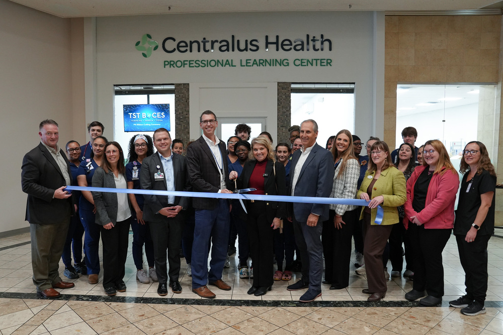 A large group of community leaders, educators, and nursing students in blue scrubs gather in front of the Centralus Health Professional Learning Center sign. In the center, TST BOCES Assistant Superintendent Cindy Walter and Centralus Health Assistant VP Dr. Michael Berlin hold large ceremonial scissors cutting a long blue ribbon stretched across the crowd.