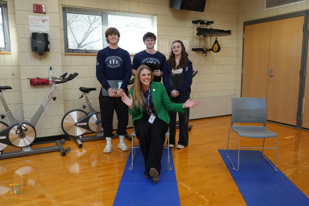 Lily Talcott sits on a mat while students stand behind her holding clipboards inside a fitness classroom.