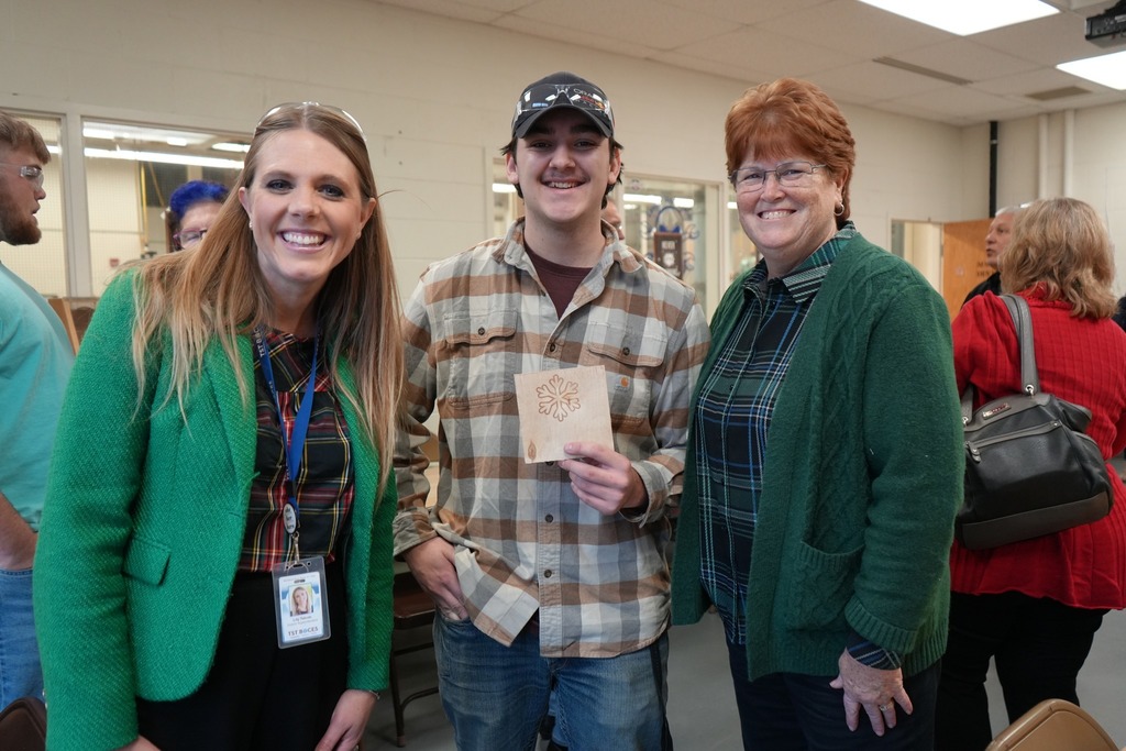A CTE Machining and Manufacturing student holds a small wooden project while standing with administrators and Board of Education members inside a classroom.