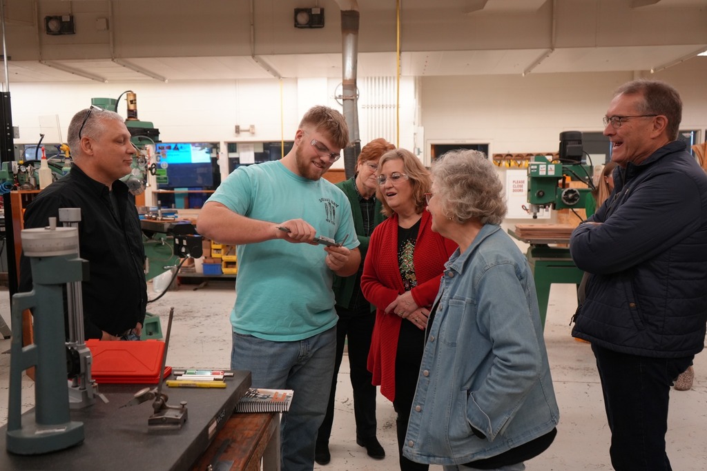 A CTE Machining and Manufacturing student demonstrates a precision measuring tool while Board of Education members and administrators observe inside a machine shop.