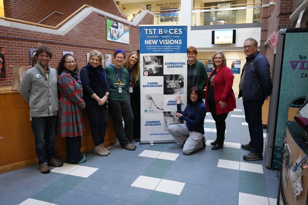 Administrators, instructors, and Board of Education members stand together near a TST BOCES New Visions banner inside a college campus building.