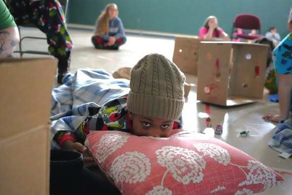 A young student lies on a pillow and blanket on the gym floor during a cozy sled-in movie, surrounded by cardboard structures and other students.