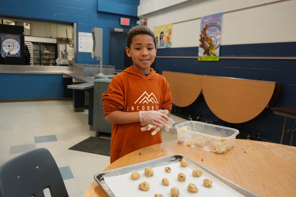 A student wearing gloves stands at a table shaping cookie dough balls on a baking sheet inside a school cafeteria.