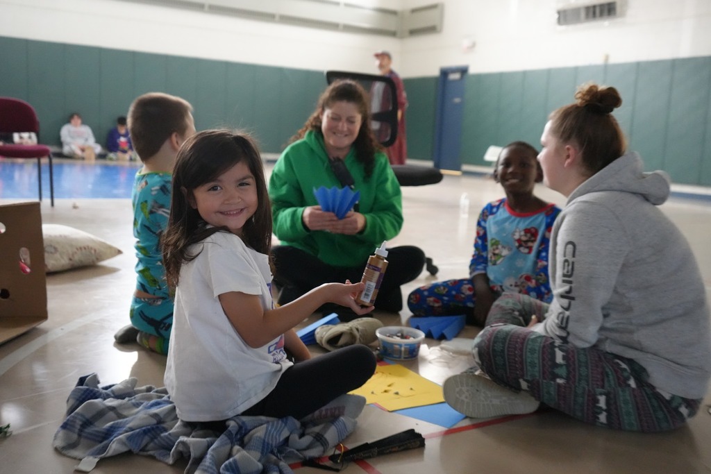 Students sit together on the gym floor working on a craft activity, with paper, supplies, and blankets spread out around them during a holiday sled-in.