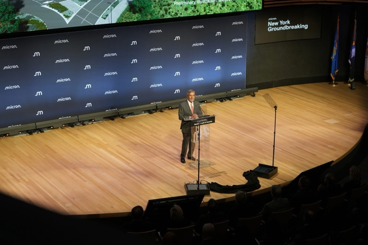 speaker, Micron CEO Sanjay MEHROTRA stands at a podium on a large stage with Micron branding behind him, addressing an audience during the Micron New York groundbreaking ceremony.