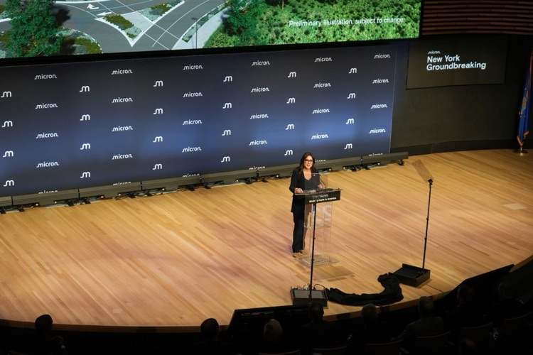 speaker at the podium on the Micron-branded stage, speaking to a seated audience during the groundbreaking event: Julie SU