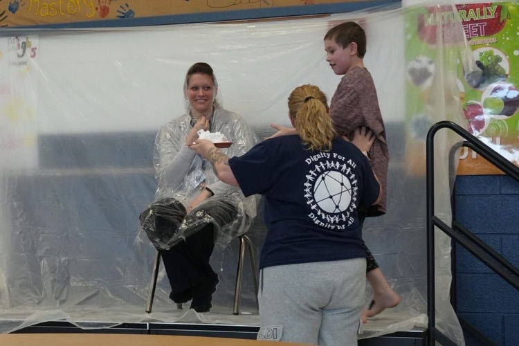 student gets on stage and makes his way toward the pie to put in staff member’s face who is sitting in a chair with a poncho over her head as she smiles 