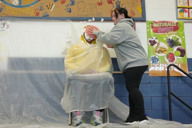 staff member sits in chair with whipped cream all over face as student smears the pie in a circular motion