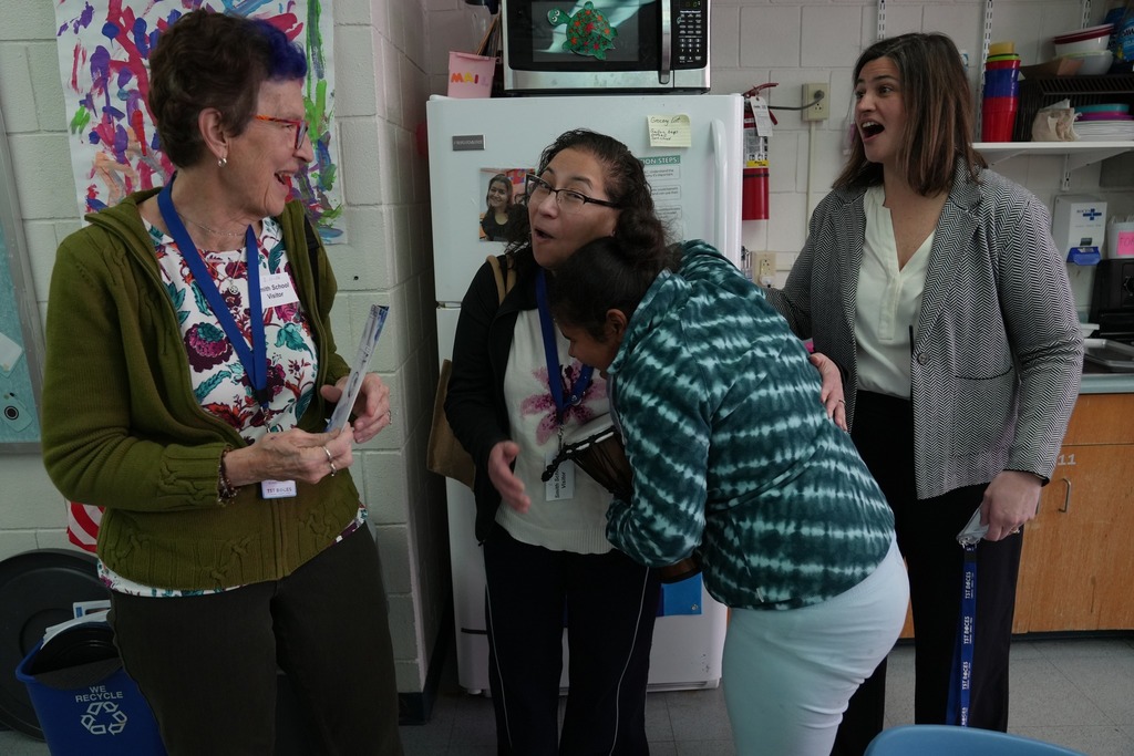 A Board of Education member smiles while speaking with a staff member and student during a classroom visit at Smith School. Another administrator stands nearby, reacting warmly as the student leans in for a hug.