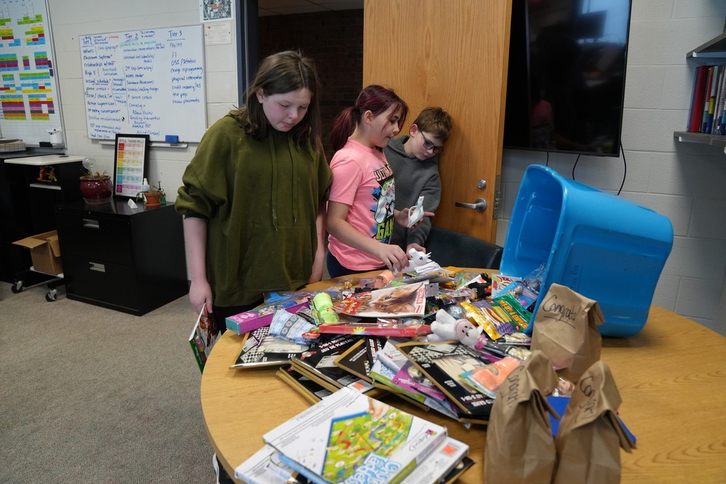 Three students stand at a table filled with small prizes, books, and reward items, choosing their selections during the High Five recognition celebration.
