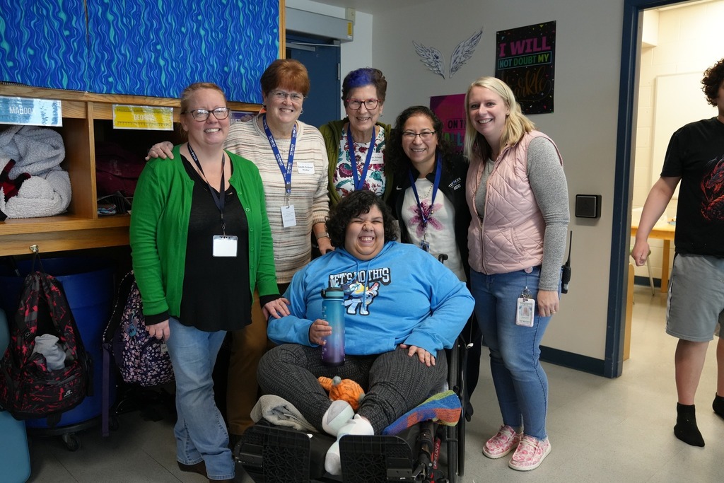 Board members and staff gather around a smiling student seated in a wheelchair in a classroom at Smith School during the tour.
