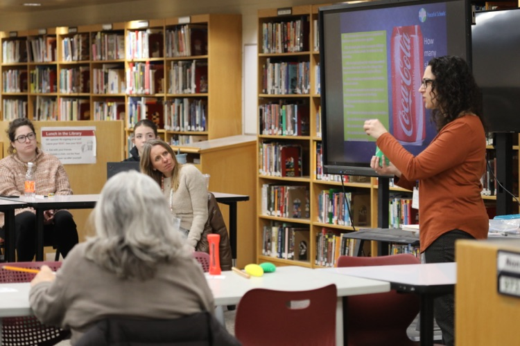  An educator presents to a small group of teachers in a library setting, using a screen display while participants listen and engage.