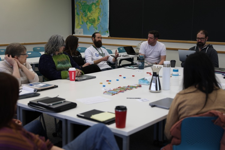  Educators sit around a table in a classroom, participating in a group discussion with materials and laptops spread out in front of them.