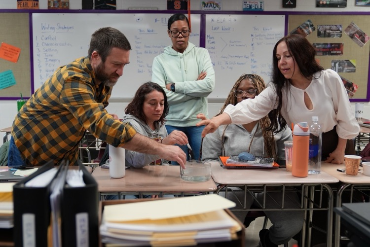  A group of educators gather around desks in a classroom, participating in a hands-on learning activity involving materials and discussion.