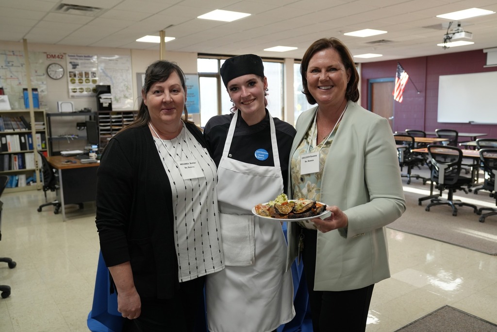 A Culinary Arts student stands between two adult guests, smiling and holding a plate of food in a classroom setting during the event.