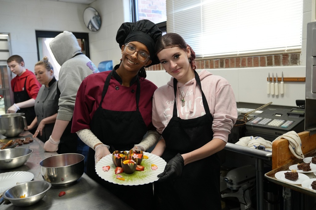 Two Culinary Arts students smile and pose together in a kitchen while presenting a plated dessert, with other students working in the background.