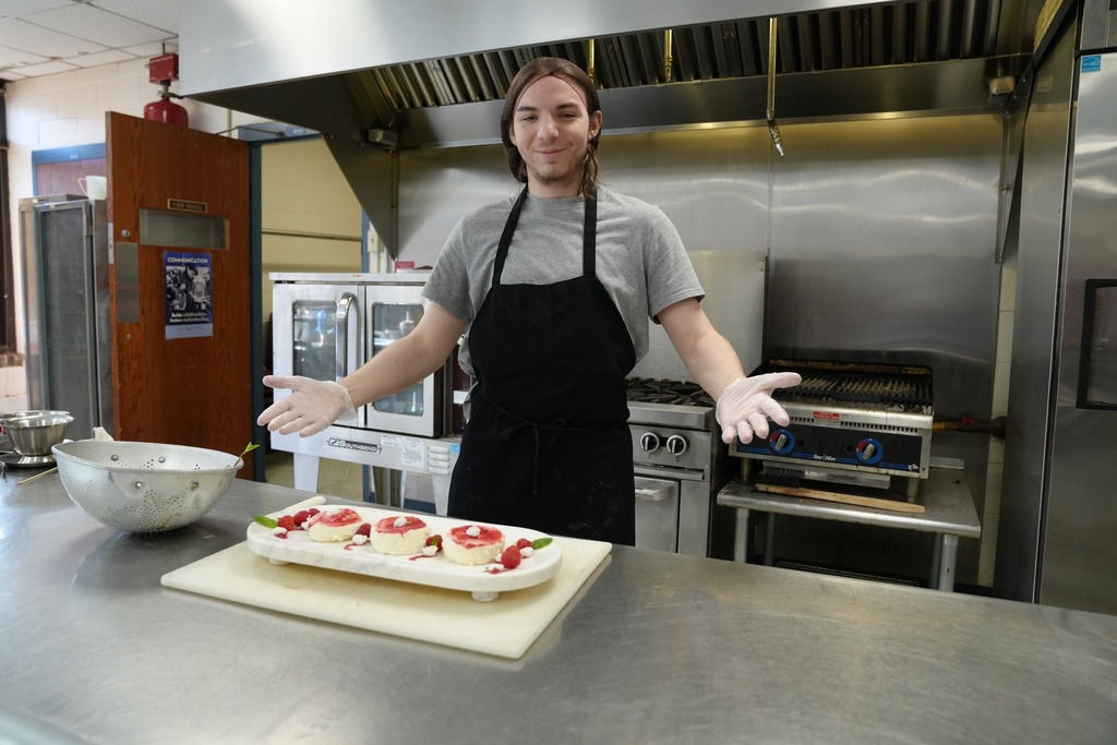 Culinary Arts student stands in a commercial kitchen presenting plated desserts, smiling with hands open in a welcoming gesture.