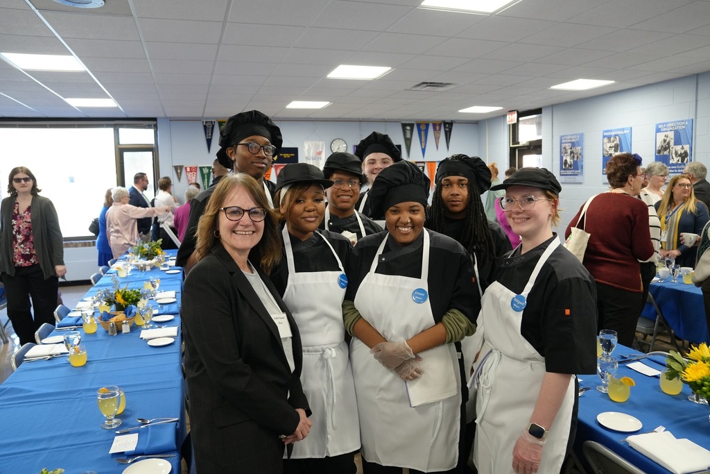 A group of Culinary Arts students in chef uniforms stand together smiling with an adult guest in a dining room set with blue tablecloths during the annual dinner event.