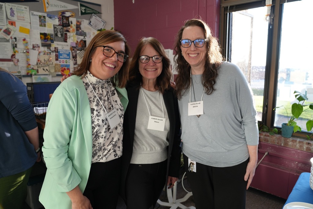 Three adults stand together smiling near a window, wearing name tags and attending the event.