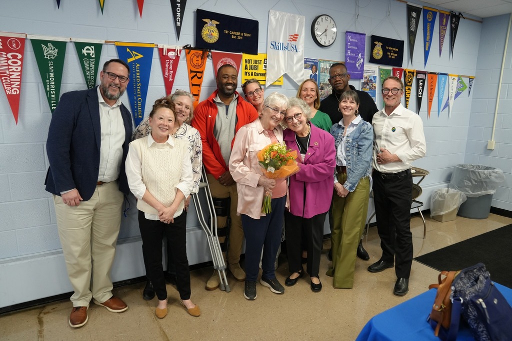 A group of adults pose together indoors, smiling as one person holds a bouquet of flowers, during an award recognition at the event.