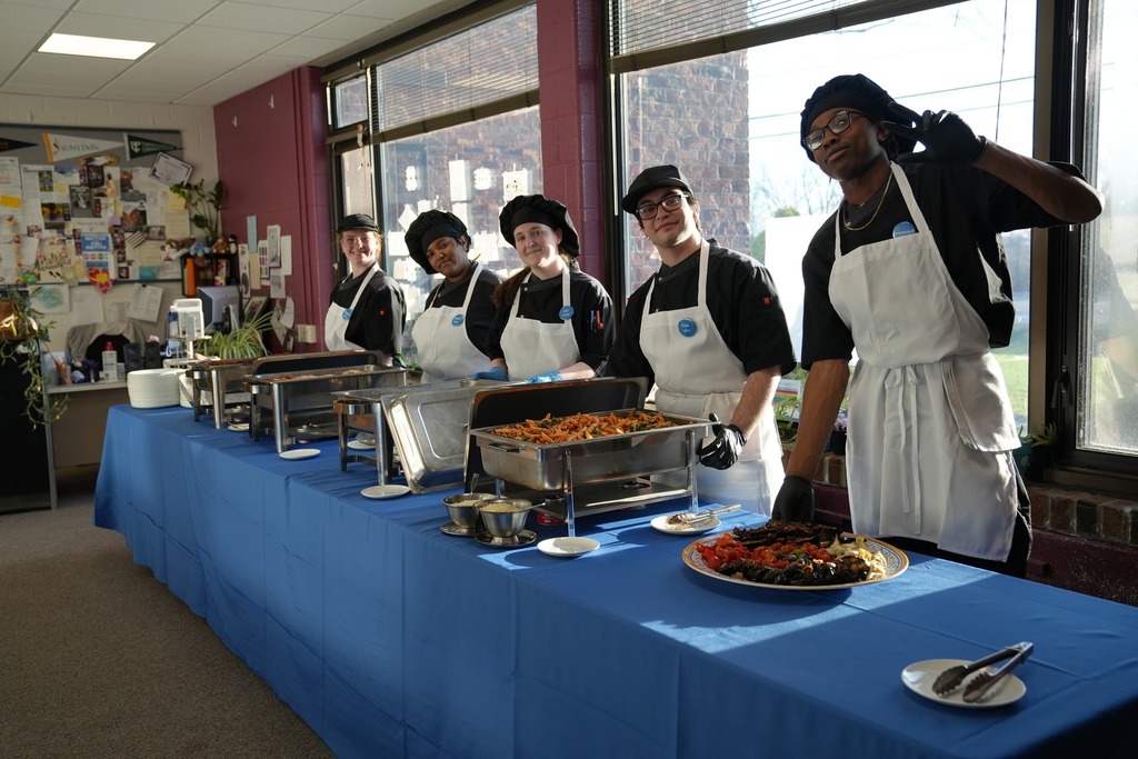 A line of Culinary Arts students in chef uniforms stand behind a buffet table filled with food, serving guests in a sunlit room.