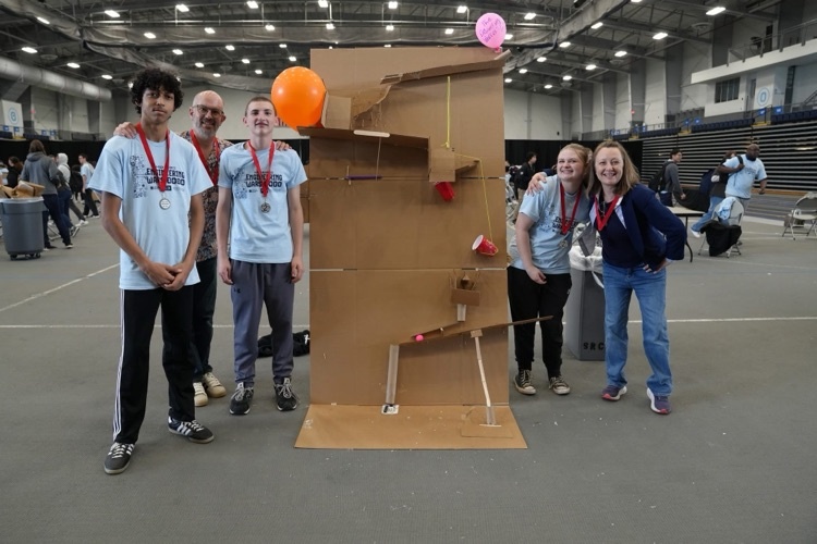  Students and an instructor stand beside a large cardboard Rube Goldberg machine with ramps, cups, and a balloon attached.