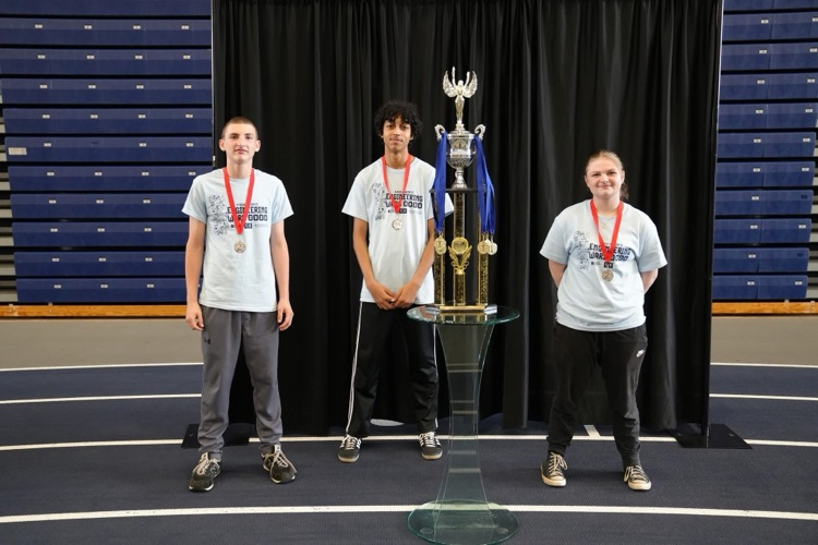  Three students wearing medals pose next to a large trophy on display in a gymnasium.