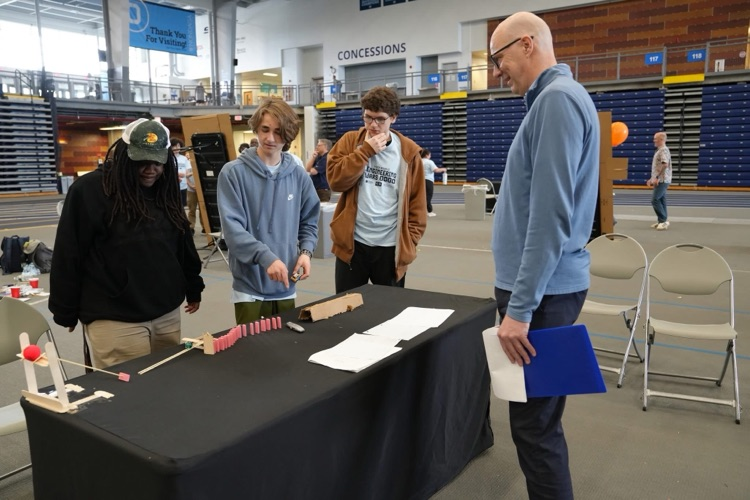  Students demonstrate a small chain-reaction setup on a table while an adult judge observes and takes notes.