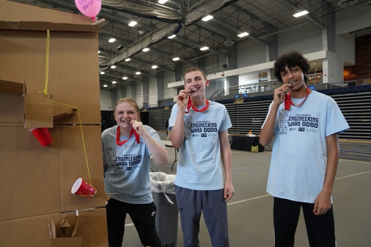           Three students smile and hold their medals while standing next to part of their cardboard Rube Goldberg machine.