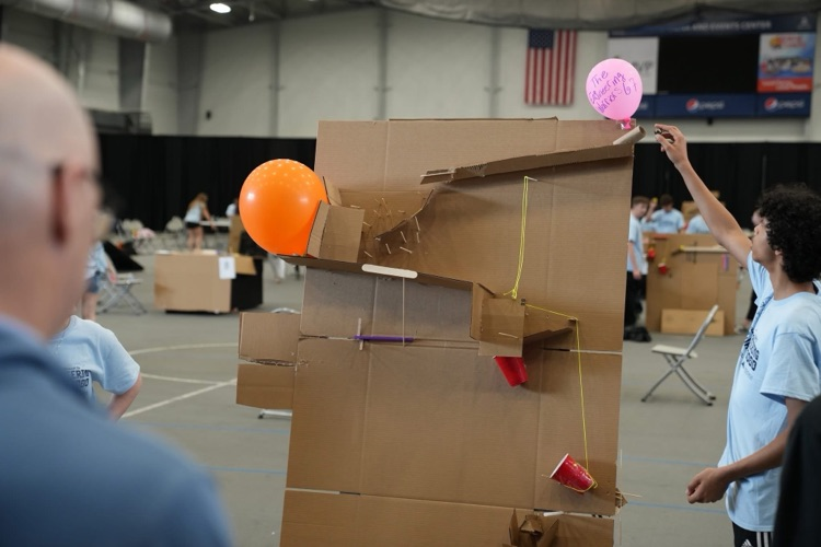A close-up of a large cardboard Rube Goldberg machine with ramps, strings, and cups as part of the design. Student is inserting marble into the top to begin the run.