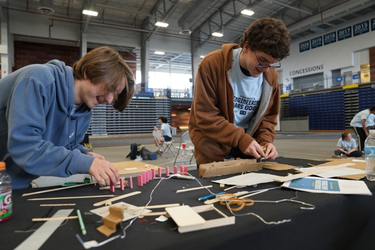 Two students work on building a chain-reaction setup with dominoes, cardboard, and string on a table.