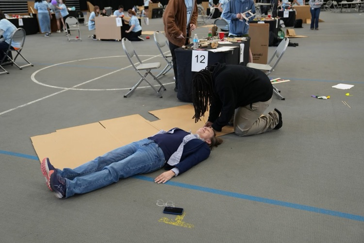 Theesfeld lies on the floor while another measures her height with a wooden strip as part of the project setup. The machine needed to be 5' long which is exactly Theesfeld's height.